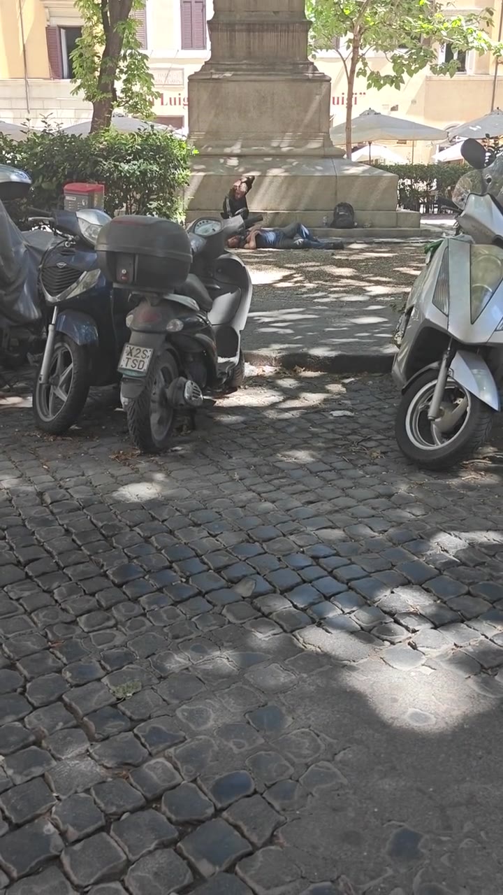 Piazza romana con sampietrini, scooter parcheggiati in fila e, alla base di un monumento in pietra con incisa la parola «Luigi», una persona sdraiata per terra all'ombra mentre un'altra le siede accanto